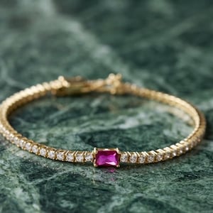 Op de afbeelding: Een goudkleurige tennisarmband met een reeks kleine, heldere, rondgeslepen stenen langs de band, met een enkele, rechthoekige, felroze edelsteen prominent in het midden van de ketting, vastgezet met een metalen sluiting.