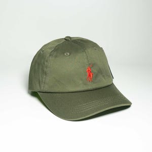 May include: An olive green baseball cap featuring a structured six-panel crown and a curved brim. The front of the cap displays a small, embroidered red logo depicting a person on a horse playing polo, centred above the brim.