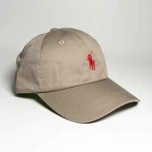 Puede incluir: Una gorra de béisbol de color tostado con una corona estructurada de seis paneles, ojales cosidos y una visera curva. La parte frontal de la gorra muestra un pequeño logotipo bordado en rojo que representa a una persona a caballo.
