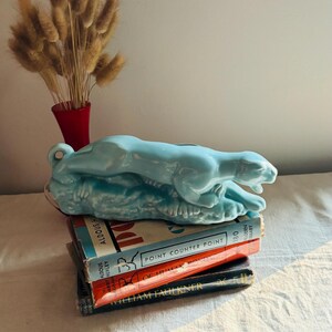 May include: A glossy light blue ceramic panther figurine in a prowling pose, resting atop a stack of three hardcover books. The books include titles by Aldous Huxley and William Faulkner, with visible text on the spines including Point Counter Point.