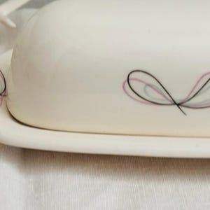 May include: A cream-colored ceramic butter dish with a domed lid and a matching base. The surface features a delicate, hand-drawn style graphic of abstract butterfly-like shapes in thin black, soft pink, and light teal lines across the exterior.