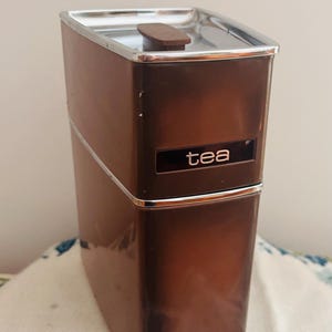 May include: A tall, rectangular brown storage canister with a metallic silver rim and a small brown handle on the lid. The canister features two distinct labels: the word tea printed near the top and the word coffee printed near the base.