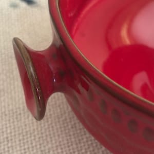May include: A bright red ceramic bowl featuring a smooth, glossy interior and a textured exterior rim with a repeating circular pattern. The bowl includes a small, protruding handle on the side with a dark brown edge finish, showcasing a vibrant color.