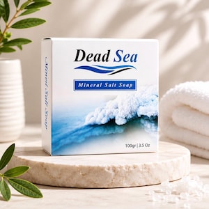 Puede incluir: Una caja blanca de jabón de sal mineral del Mar Muerto se muestra sobre un pedestal de piedra. La caja presenta las palabras "Dead Sea" en negro, con un diseño de onda azul y "Mineral Salt Soap" en un rectángulo azul. La caja también indica 100gr.