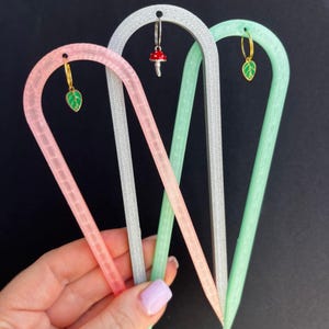 May include: Three U-shaped plant trellises in pink, silver, and light green, each featuring a small dangling charm. The pink and green trellises have green leaf charms, while the silver one has a red mushroom charm. The text PLANTCEBO is visible.