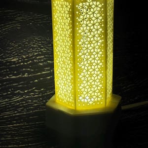 May include: A hexagonal lamp with a yellow, intricate, laser-cut geometric pattern on its sides. The lamp features a solid base and a matching top rim, emitting a warm, bright glow from within that highlights the detailed lattice structure.