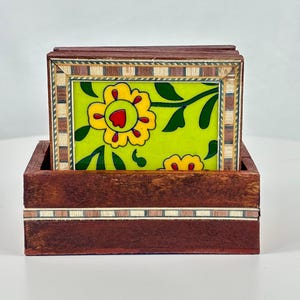 May include: A set of rectangular coasters stored in a dark brown wooden box. Each coaster features a bright lime green background with a yellow flower design, red center, and dark green leaves, framed by a border of alternating light and dark wood.