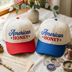 Puede incluir: Dos gorras de béisbol con coronas color hueso y texto bordado en azul que dice American y en rojo HONEY con estrellas azules. Una gorra tiene visera y botón rojos, mientras que la otra tiene visera y botón de color azul.