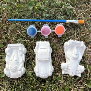 Può includere: Un set di tre figurine di auto in ceramica bianca non dipinte, un pennello con manico blu e setole arancioni, e una striscia di tre piccoli vasetti di vernice in plastica contenenti vernice blu, rosa e arancione.
