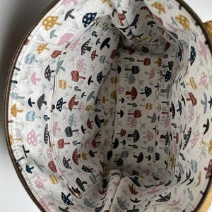 May include: A round fabric bag interior featuring a cream-colored lining with a repeating pattern of small mushrooms in shades of mustard yellow, navy blue, dusty pink, and dark red, including an integrated interior pocket with a zipper.
