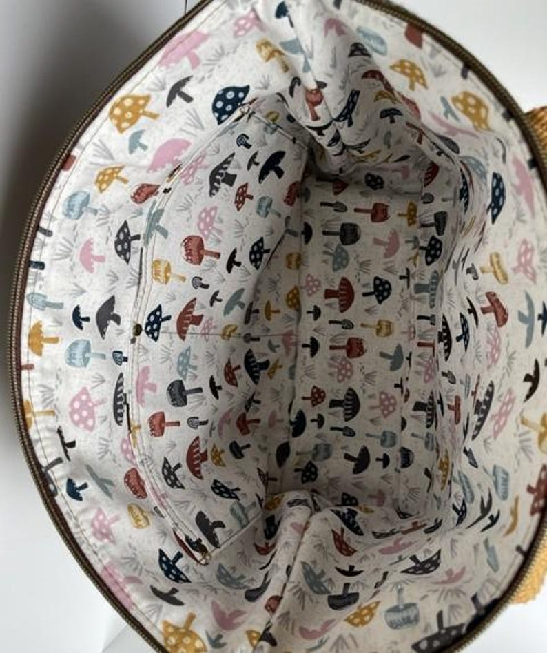 May include: A round fabric bag interior featuring a cream-colored lining with a repeating pattern of small mushrooms in shades of mustard yellow, navy blue, dusty pink, and dark red, including an integrated interior pocket with a zipper.