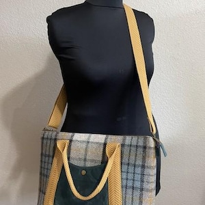 May include: A rectangular tote bag featuring a grey, blue, and yellow plaid pattern. The bag includes a dark green front pocket with a single brass-colored snap closure, yellow herringbone-patterned webbing handles, and a matching yellow shoulder strap.