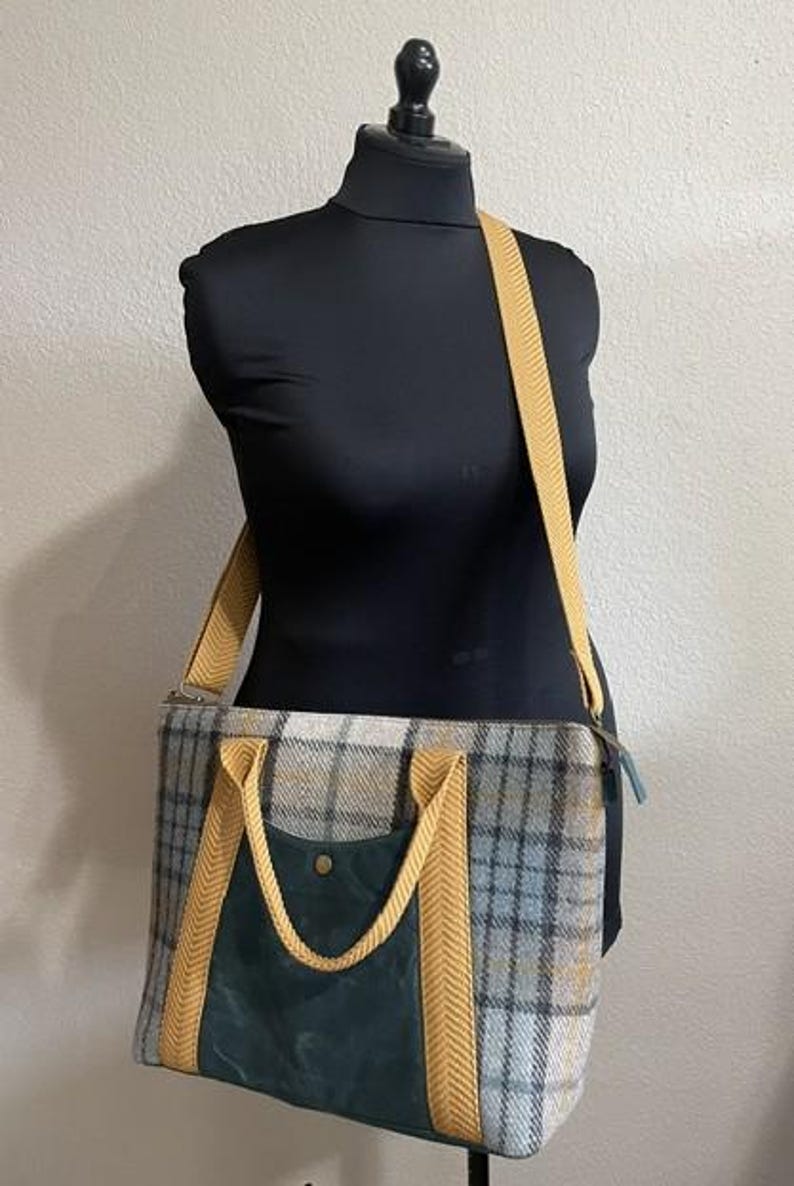 May include: A rectangular tote bag featuring a grey, blue, and yellow plaid pattern. The bag includes a dark green front pocket with a single brass-colored snap closure, yellow herringbone-patterned webbing handles, and a matching yellow shoulder strap.