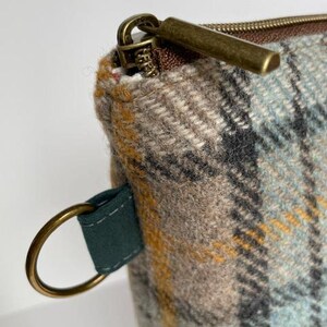 May include: A zippered pouch made of woven wool fabric featuring a plaid pattern in shades of light blue, tan, gray, and mustard yellow. The pouch includes a dark green fabric tab with a circular brass ring and a brass-toned metal zipper pull.
