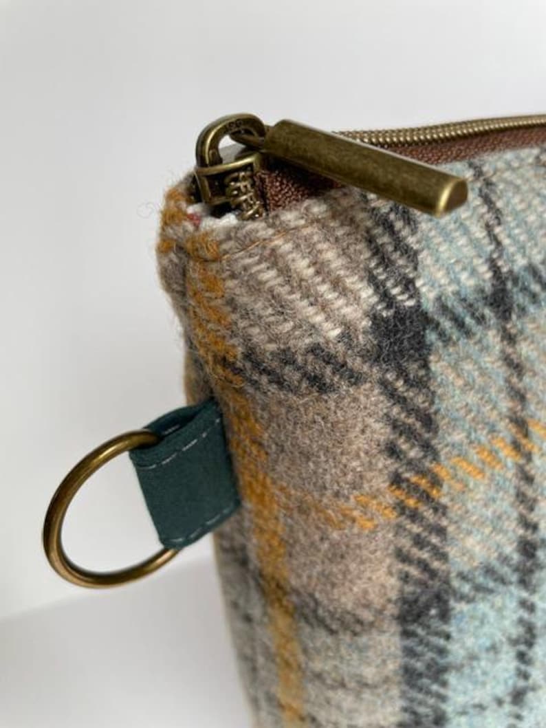 May include: A zippered pouch made of woven wool fabric featuring a plaid pattern in shades of light blue, tan, gray, and mustard yellow. The pouch includes a dark green fabric tab with a circular brass ring and a brass-toned metal zipper pull.