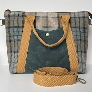 May include: A rectangular tote bag with a plaid pattern in shades of gray, blue, and tan. The bag features a dark teal front pocket and tan-colored handles and a shoulder strap. The bag has a zipper closure and metal hardware.