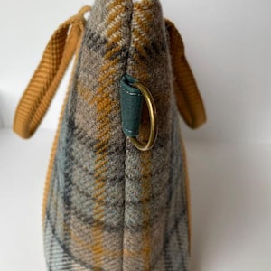 May include: A side view of a tote bag made of a grey, tan, and mustard yellow plaid fabric. The bag features mustard yellow woven handles, a dark teal fabric tab with a brass-toned metal D-ring, and a brass-toned metal zipper at the top.