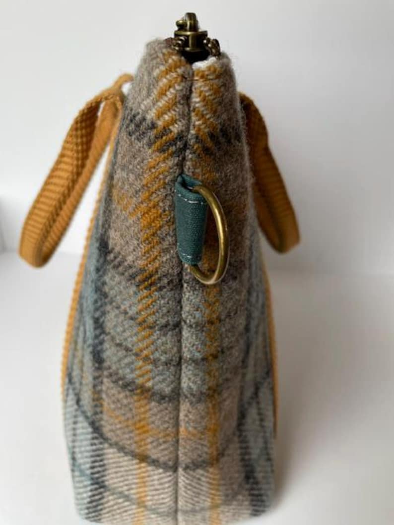 May include: A side view of a tote bag made of a grey, tan, and mustard yellow plaid fabric. The bag features mustard yellow woven handles, a dark teal fabric tab with a brass-toned metal D-ring, and a brass-toned metal zipper at the top.