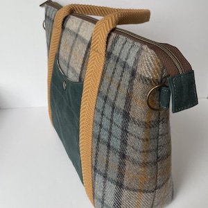 May include: A rectangular tote bag featuring a grey, blue, and yellow plaid wool pattern. It includes a dark green front pocket with a metal snap, mustard yellow herringbone webbing handles, a brown zipper closure, and a brass D-ring on the side.