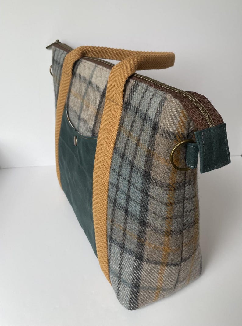May include: A rectangular tote bag featuring a grey, blue, and yellow plaid wool pattern. It includes a dark green front pocket with a metal snap, mustard yellow herringbone webbing handles, a brown zipper closure, and a brass D-ring on the side.