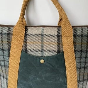 May include: A tote bag featuring a grey, blue, and tan plaid pattern with a dark teal front pocket secured by a single brass-colored snap button. The bag includes two thick, textured mustard yellow woven fabric handles attached to the front.