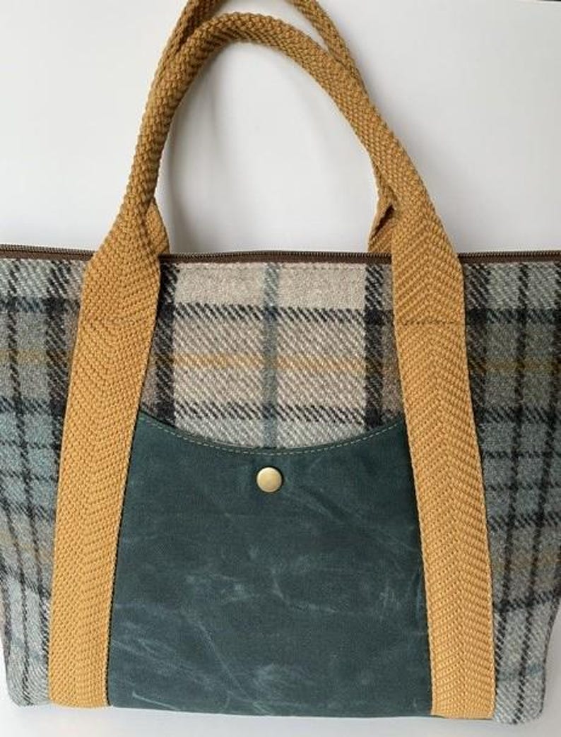 May include: A tote bag featuring a grey, blue, and tan plaid pattern with a dark teal front pocket secured by a single brass-colored snap button. The bag includes two thick, textured mustard yellow woven fabric handles attached to the front.