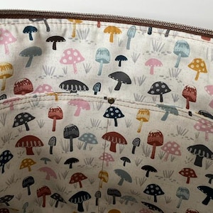 May include: The interior of a bag featuring a cream-colored fabric lining with a repeating pattern of mushrooms in various colors including pink, blue, yellow, and dark brown. A horizontal pocket is stitched across the middle with a brown zipper top.