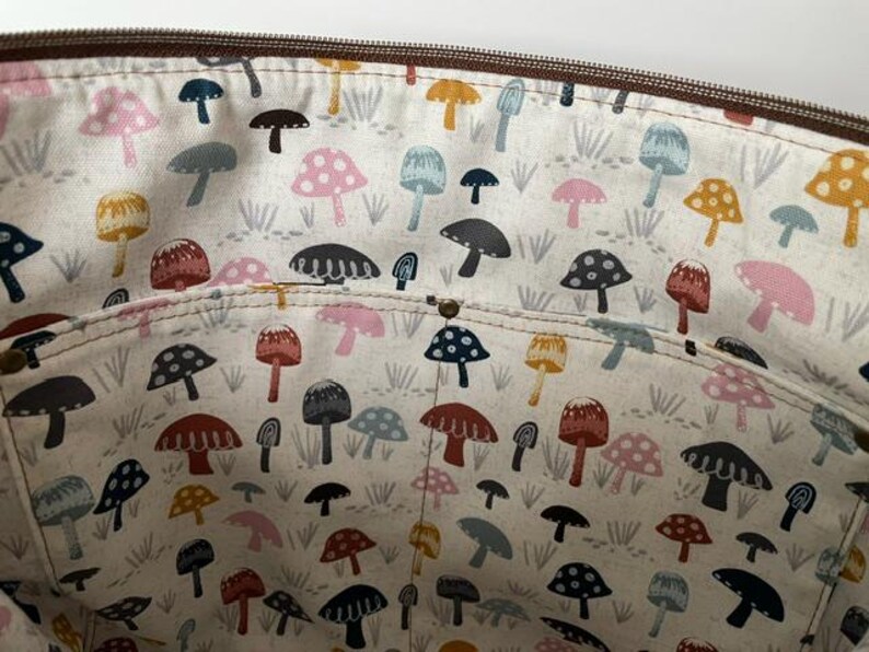 May include: The interior of a bag featuring a cream-colored fabric lining with a repeating pattern of mushrooms in various colors including pink, blue, yellow, and dark brown. A horizontal pocket is stitched across the middle with a brown zipper top.