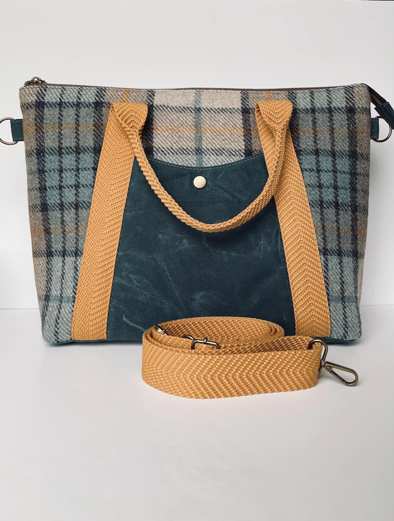 May include: A rectangular handbag featuring a blue, gray, and tan plaid pattern with a central dark teal pocket secured by a gold-tone snap. The bag includes mustard yellow herringbone webbing handles and a matching detachable shoulder strap.