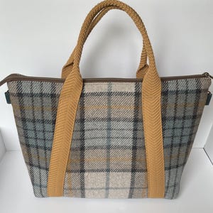 May include: A tote bag featuring a grey, beige, and blue plaid pattern with a brown zipper closure. The bag has two thick, textured mustard yellow fabric handles that extend down the front and back panels, providing a sturdy grip for carrying.