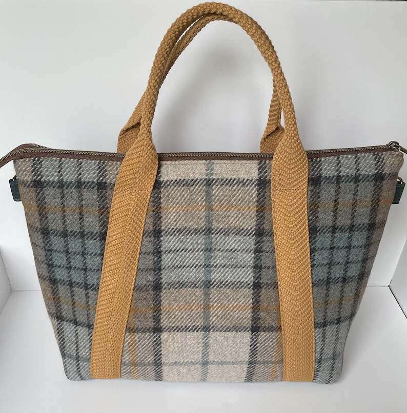 May include: A tote bag featuring a grey, beige, and blue plaid pattern with a brown zipper closure. The bag has two thick, textured mustard yellow fabric handles that extend down the front and back panels, providing a sturdy grip for carrying.