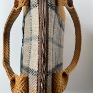 May include: A rectangular bag featuring a beige, gray, and mustard yellow plaid pattern. The bag has a brown central zipper closure and two mustard yellow woven fabric handles attached to the sides. A dark green accent panel is visible on the side.