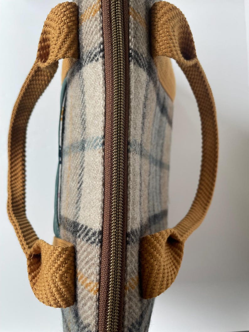 May include: A rectangular bag featuring a beige, gray, and mustard yellow plaid pattern. The bag has a brown central zipper closure and two mustard yellow woven fabric handles attached to the sides. A dark green accent panel is visible on the side.