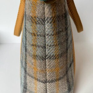 May include: A tote bag featuring a plaid pattern in shades of mustard yellow, light blue, and grey. The bag has mustard yellow woven fabric handles and a dark green fabric tab with a circular bronze-toned metal ring attached to the top edge.