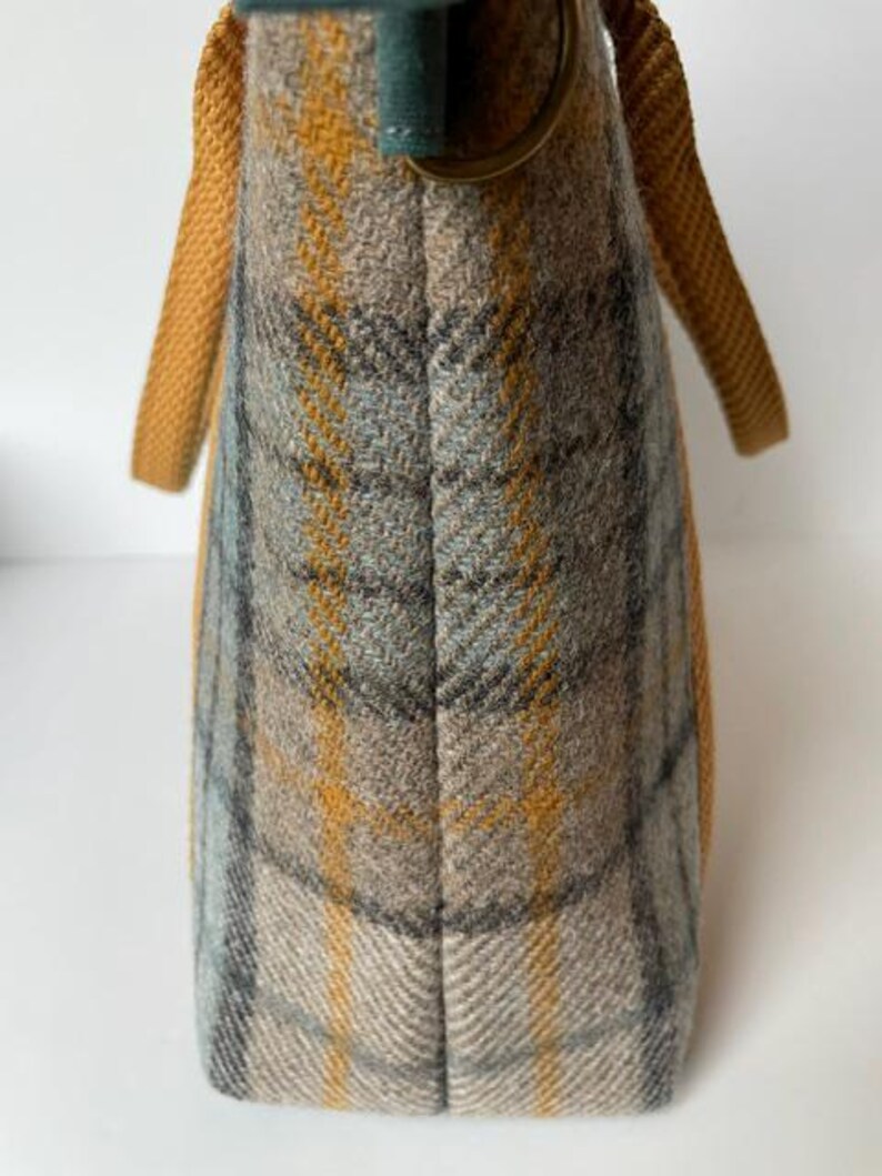 May include: A tote bag featuring a plaid pattern in shades of mustard yellow, light blue, and grey. The bag has mustard yellow woven fabric handles and a dark green fabric tab with a circular bronze-toned metal ring attached to the top edge.
