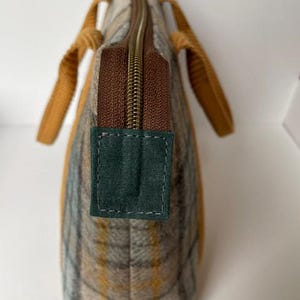 May include: A tote bag featuring a plaid pattern in shades of tan, gray, and yellow. The bag has a brown zipper closure with a dark green fabric tab at the end. Two mustard yellow textured handles are attached to the sides of the bag.