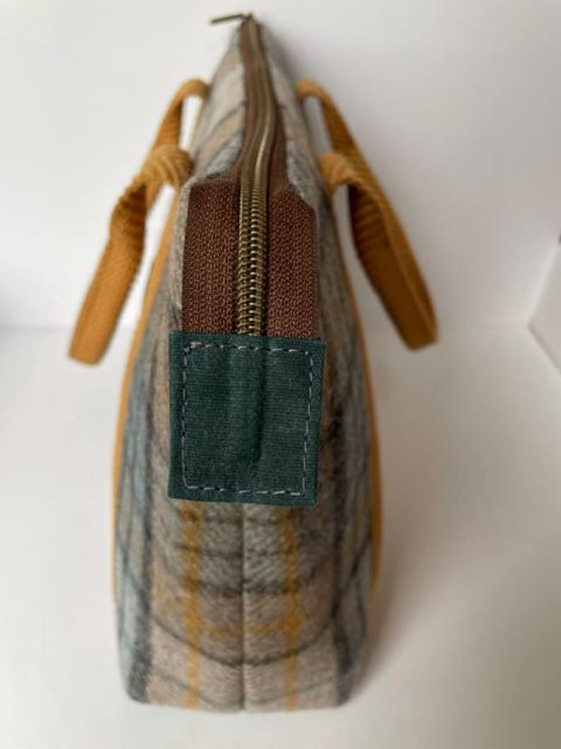 May include: A tote bag featuring a plaid pattern in shades of tan, gray, and yellow. The bag has a brown zipper closure with a dark green fabric tab at the end. Two mustard yellow textured handles are attached to the sides of the bag.
