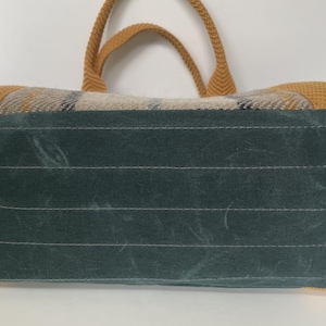 May include: A rectangular bag featuring a dark teal base with four brass-toned metal feet. The upper portion displays a woven plaid pattern in shades of tan, gray, and black, complemented by mustard yellow woven handles and side accents.
