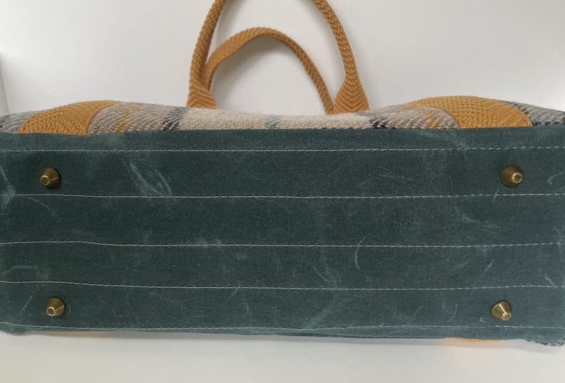 May include: A rectangular bag featuring a dark teal base with four brass-toned metal feet. The upper portion displays a woven plaid pattern in shades of tan, gray, and black, complemented by mustard yellow woven handles and side accents.
