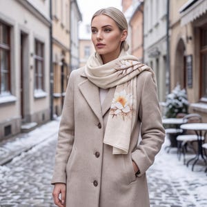 May include: A beige wool coat with a notched lapel and three front buttons, paired with a cream-colored scarf featuring a printed white and yellow lotus flower design with brown accents, worn around the neck with one end draped over the shoulder.