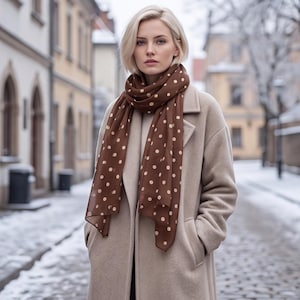 May include: A brown scarf featuring a repeating pattern of small, light-colored polka dots, worn over a beige wool-blend coat with a notched lapel and deep front pockets. The scarf is draped loosely around the neck, with ends hanging down.