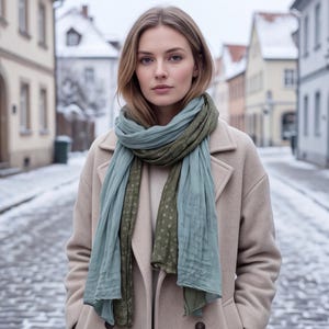 May include: A beige wool coat worn with a long, lightweight scarf featuring a dual-tone design. The scarf has a soft, crinkled texture with one side in a muted sage green and the other in a darker olive green, accented by a subtle, repeating pattern.