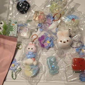 May include: A selection of small, colourful charms and keyrings, individually packaged in clear plastic. The charms showcase diverse designs, including cartoon characters, animals, and geometric shapes, with glitter and vibrant colours.