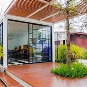 May include: A modern outdoor structure with a wooden deck and a white frame. Large glass sliding doors reveal an interior with a checkered floor. The structure is partially shaded by a slatted wooden roof. Green plants and trees surround the building.