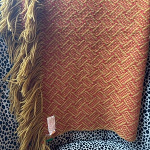 May include: A woven throw blanket featuring a geometric pattern in shades of orange and mustard yellow. The blanket has long, thick mustard yellow fringe along one side and a small white rectangular fabric tag attached near the bottom edge.