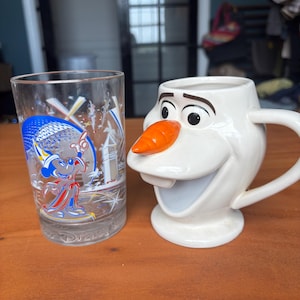 May include: A clear glass tumbler featuring a colorful graphic of Mickey Mouse in a wizard hat and a white ceramic mug sculpted in the shape of the character Olaf with a prominent orange carrot nose and a wide, open-mouthed expression.