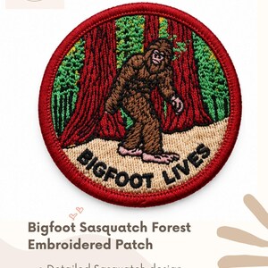 May include: A circular embroidered patch featuring a brown Bigfoot figure walking through a forest of tall red trees and green foliage. The patch has a red border and the text BIGFOOT LIVES embroidered in black at the bottom on a tan background.