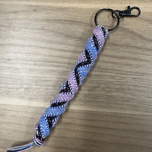 以下が含まれることがあります： 青、紫、黒、白の色合いの編み込みキーホルダー。キーホルダーには黒い金属リングとロブスタークラスプが付いています。編み込み部分は約15cmです。