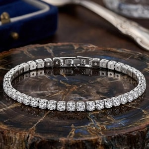 Può includere: Un bracciale tennis color argento caratterizzato da una fila continua di pietre rotonde, trasparenti e sfaccettate, incastonate in maglie metalliche quadrate. Il bracciale include una chiusura a cassetta sicura con finitura metallica lucida.