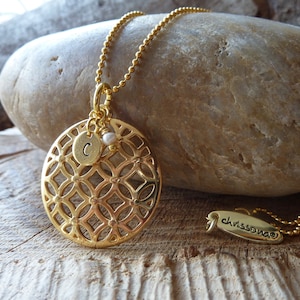 Könnte beinhalten: Goldkette mit einem runden Anhänger mit geometrischem Muster und einem kleinen Anhänger mit dem Buchstaben 'C'. Der Anhänger ist mit einer kleinen Perle verziert. Die Kette liegt auf einem Stein und einem Etikett mit der Aufschrift "chrisswong".